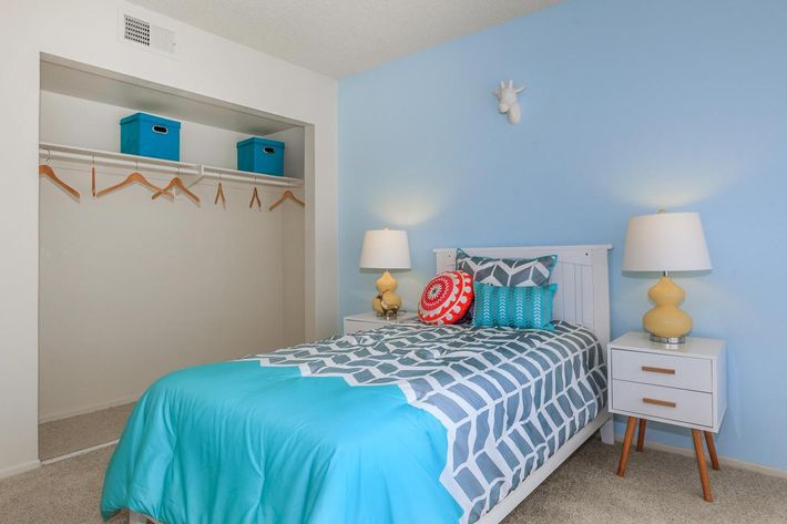 A cozy bedroom featuring a twin bed with a turquoise quilt, decorative pillows, and matching bedside tables with yellow lamps. A minimalist white closet with hanging space is visible, along with blue storage boxes above. The walls are painted light blue, creating a calm atmosphere.