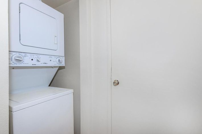 A stacked washer and dryer unit located in a small laundry space next to a closed white door. The walls are painted a light color, and the overall setting appears clean and well-maintained.