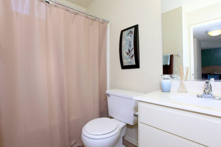 A clean bathroom featuring a light pink shower curtain, a toilet, and a white sink with a modern faucet. The sink area includes decorative items like a small jar and reed diffuser. A framed artwork hangs on the wall, and a mirror reflects part of the room's interior. Natural light illuminates the space.