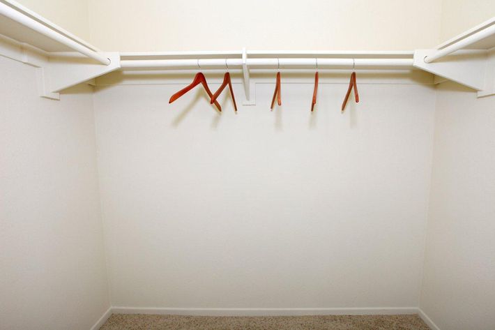An empty closet with beige walls and carpeted floor, featuring a white hanging rod with several orange hangers. There are no clothes or items hanging from the hangers, creating a spacious and minimalistic appearance.