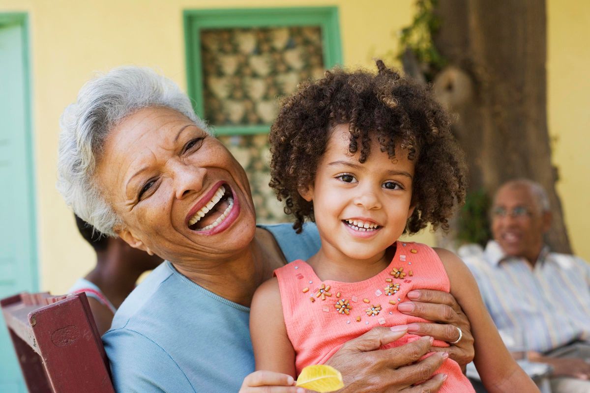 An elderly woman with short gray hair and a bright smile holds a young girl with curly hair, who is also smiling. They are outdoors, surrounded by vibrant colors. The woman is wearing a light blue shirt, while the girl is in a pink dress adorned with embellishments. A blurred figure of another person sits in the background.