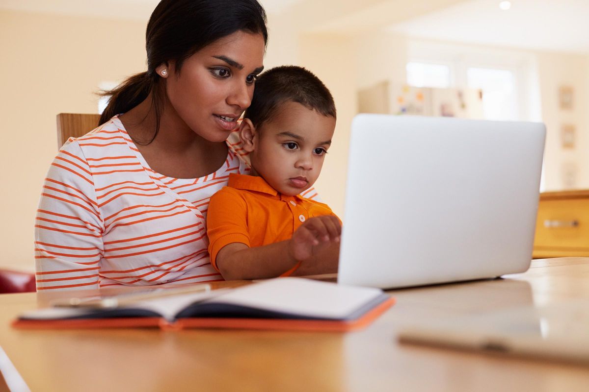A mother and her young son sit closely together at a table, engaged in looking at a laptop screen. The mother, wearing a striped shirt, appears focused while the boy, dressed in an orange shirt, curiously reaches towards the laptop. A notebook is open on the table in front of them.