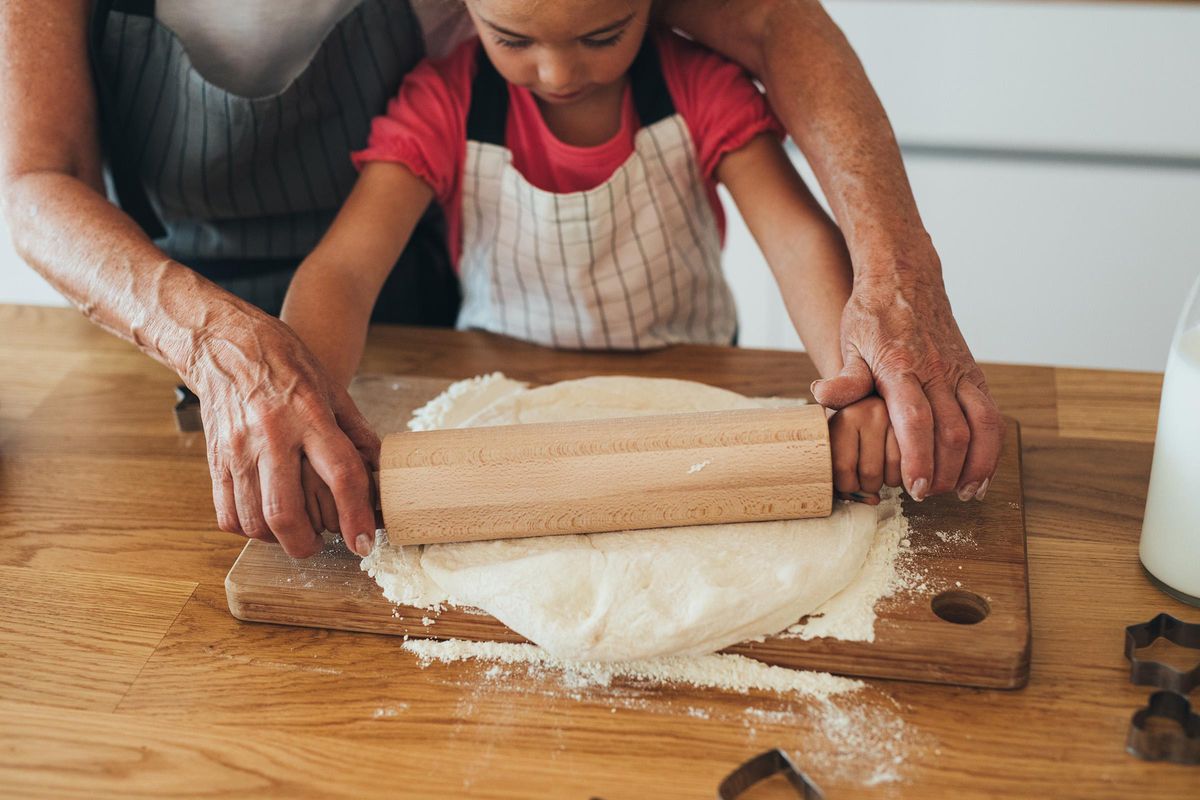 A close-up of a child's hands and an adult's hands using a rolling pin to flatten dough on a wooden surface. Both are wearing aprons, and flour is scattered around. The scene captures a warm baking moment, highlighting the bond between the adult and child.