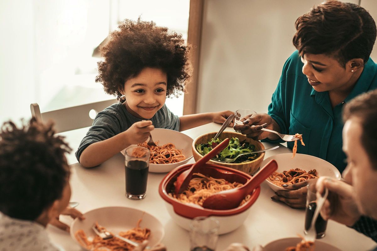 A family enjoying a meal together at a dining table. A child with curly hair smiles while eating pasta, surrounded by bowls of spaghetti and salad. Two adults engage in conversation, creating a warm, joyful atmosphere of togetherness during the meal.