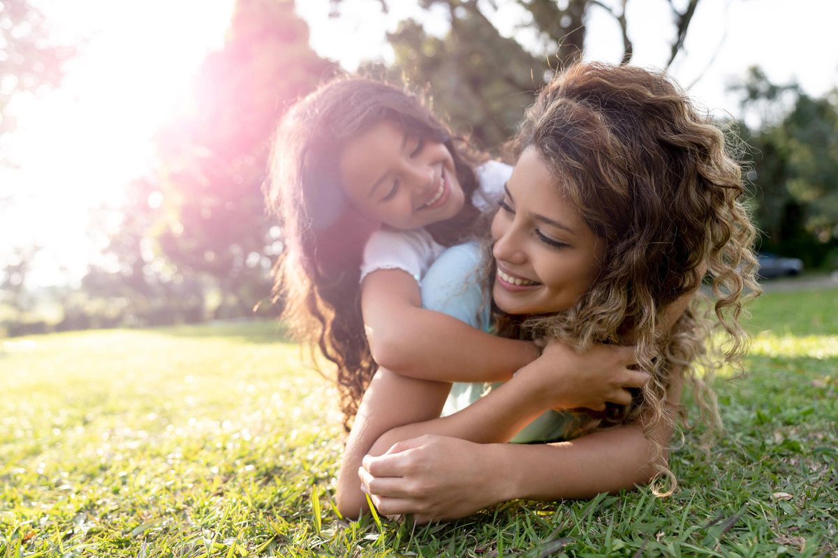 A joyful mother and daughter embrace on a grassy field during a sunny day. The little girl is playfully hugging her mother from behind, both sharing smiles and laughter, surrounded by nature and soft sunlight, creating a warm and loving atmosphere.