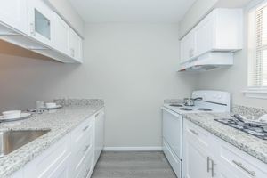 A modern kitchen featuring white cabinets and countertops, stainless steel appliances including an oven and stovetop, and a single sink. The walls are painted a light color, and there are dishes set on the counter. Large windows allow natural light into the space.