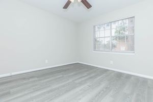 A blank, unfurnished room with light gray wooden flooring and pale gray walls. There is a ceiling fan with wooden blades, and a window with blinds allowing soft natural light to enter. The room has no furniture or decor, creating a minimalist and spacious atmosphere.