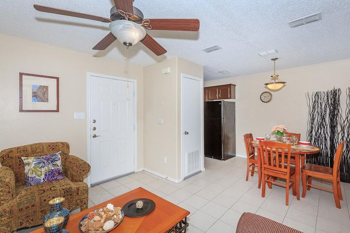 Interior view of a cozy living space featuring a comfortable brown sofa with patterned cushions, a wooden coffee table adorned with decorative items, and a dining area with a round table set for four. A ceiling fan hangs above, and there are light-colored walls with simple decor. The entrance door is visible on the left.