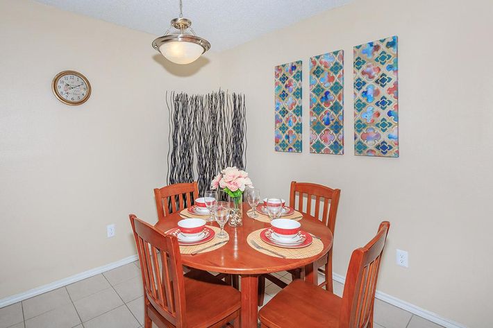 A cozy dining area featuring a round wooden table set for four, with plates, glasses, and a flower centerpiece. The walls are a warm beige, accented by three colorful decorative art pieces. A clock is mounted on the wall, and there are decorative tall grass arrangements nearby.