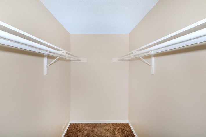 A spacious empty closet featuring two white hanging rods supported by brackets on either side. The walls are painted a neutral color, and the floor is carpeted in a light brown hue. The ceiling is plain and blue, creating a bright and airy atmosphere.