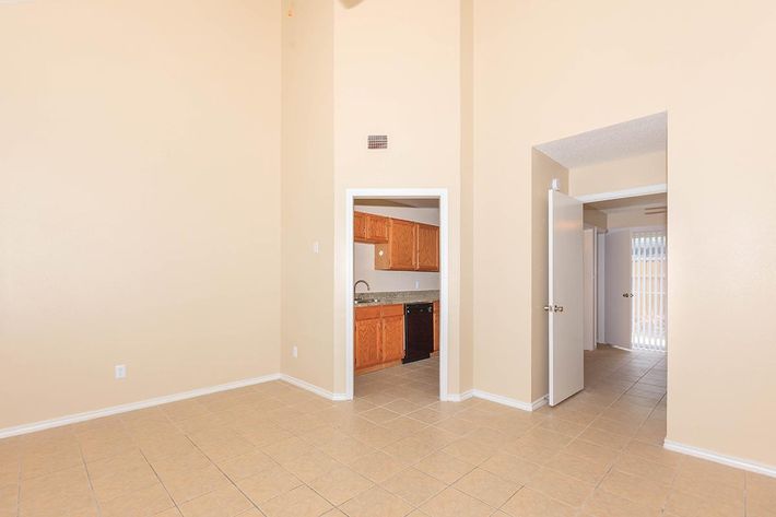 A spacious room with beige walls and tiled flooring, featuring an open doorway leading to a kitchen area with wooden cabinets. A door on the right leads outside, and there is ample natural light coming through the adjacent window. The high ceiling adds to the room's openness and brightness.