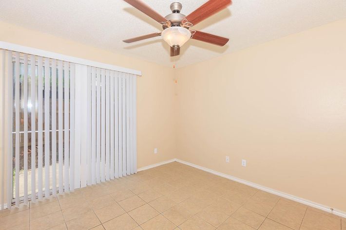 A spacious, empty room with beige walls and tile flooring. A ceiling fan with a light fixture is mounted on the ceiling. On one side, there's a set of vertical blinds covering a window that leads to the outdoors. The room has a clean, minimalist feel, suitable for various uses.