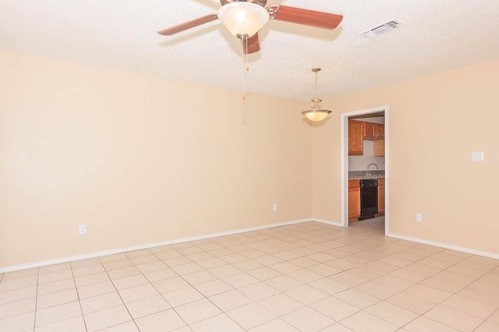 A spacious, empty room with beige walls and tiled flooring. There is a ceiling fan and a light fixture hanging from the ceiling. An open doorway leads to a kitchen area with wooden cabinets visible in the background. The overall atmosphere is bright and inviting, suitable for various interior designs.