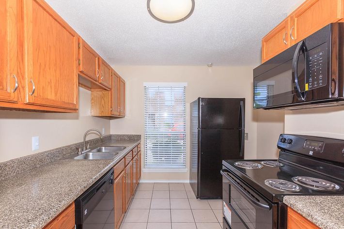 A modern kitchen featuring wooden cabinets, a granite countertop, a double sink, and essential appliances including a black refrigerator, a microwave, and an electric stove. Natural light enters through a window with blinds, illuminating the tiled floor.