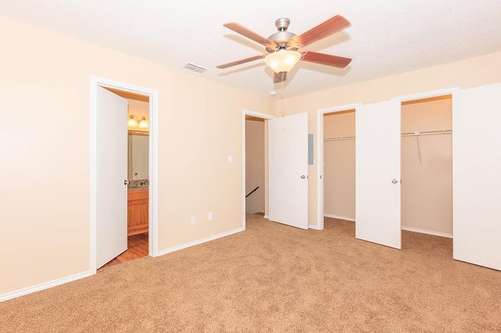 A spacious, empty bedroom featuring a ceiling fan, light tan walls, and beige carpet. There are two white doors and an open closet with a hanging rod. A bathroom is visible through one of the doors, offering a glimpse of fixtures. The room is bright and ready for furnishing.