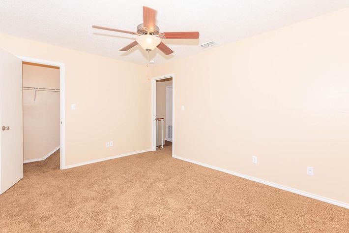 A spacious, empty room with beige walls and brown carpeting. A ceiling fan with light fixture is mounted in the center. There are two doorways leading to adjacent rooms, and a small closet is visible on the left wall. The overall atmosphere is bright and inviting, ideal for personal customization.