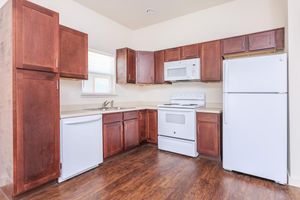 A modern kitchen featuring wooden cabinetry with a light brown finish, a white refrigerator, a white microwave above the stove, and a white oven. The countertops are beige, and there is a kitchen sink underneath a window that lets in natural light. The flooring is a dark wood laminate.