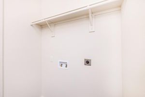 Empty laundry room with a white wall, featuring a shelf mounted above and two utility connections for a washer. There is a power outlet on the right side of the wall, and the overall color scheme is neutral, highlighting cleanliness and simplicity.