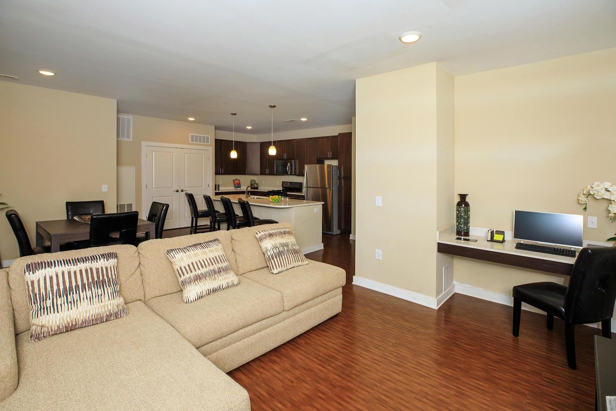 A spacious living area featuring a beige sectional sofa with patterned cushions, a dining table with black chairs, and a modern kitchen in the background. The room has warm wood flooring and neutral-colored walls, with a small workspace against one wall, complete with a computer and decorative elements.