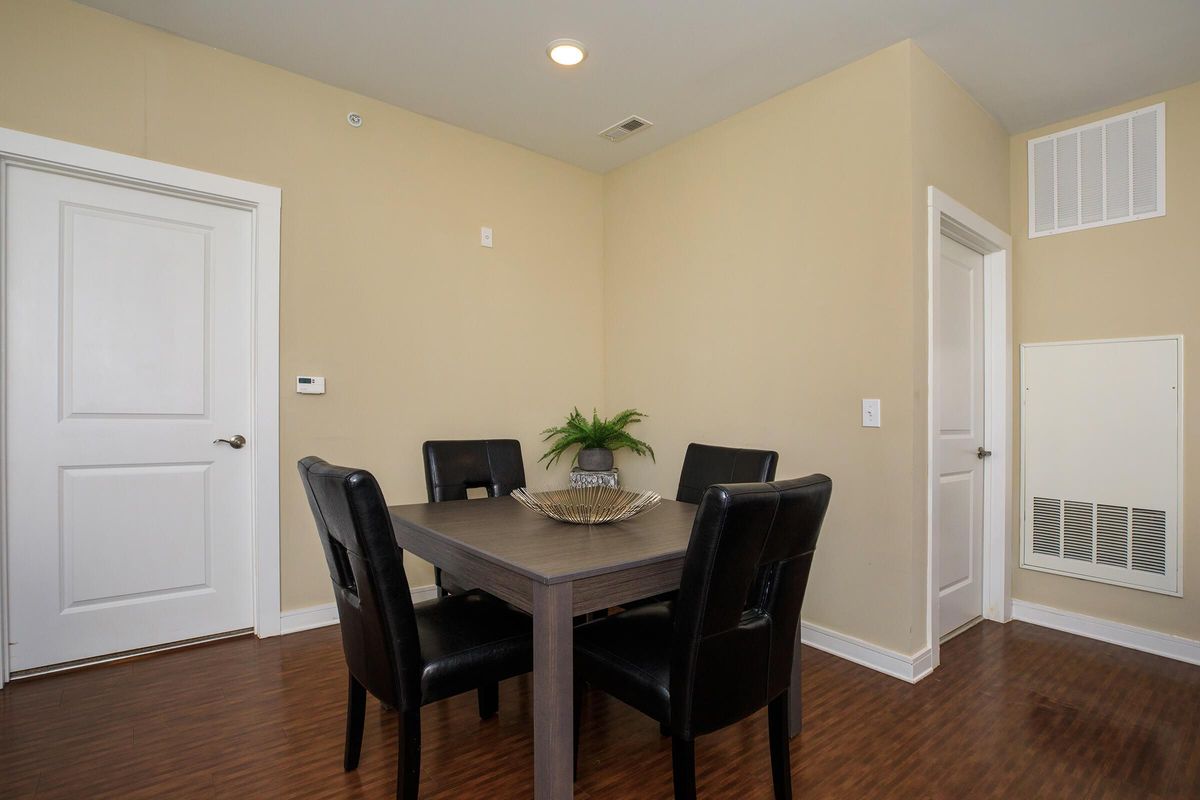 A small dining area featuring a square wooden table surrounded by four black leather chairs. The walls are painted a light beige, and there's a potted plant in the center of the table. A door is visible on the left, and the room has wood laminate flooring. An air vent is seen on the right wall.