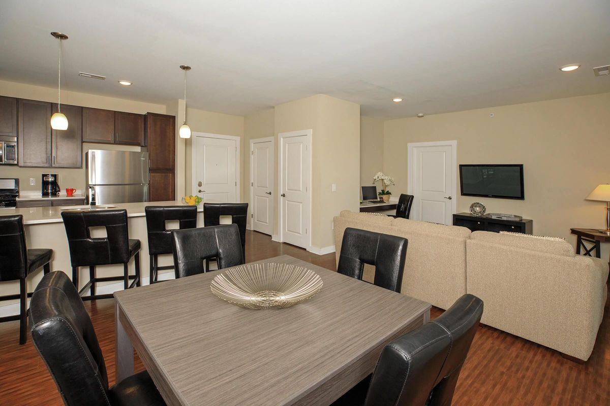 Modern living room and kitchen area featuring a dining table with black chairs, a beige sofa, and a kitchen bar with stools. The kitchen includes stainless steel appliances. Light walls and flooring create a spacious and inviting ambiance. Functional layout with a television and workspace visible in the background.