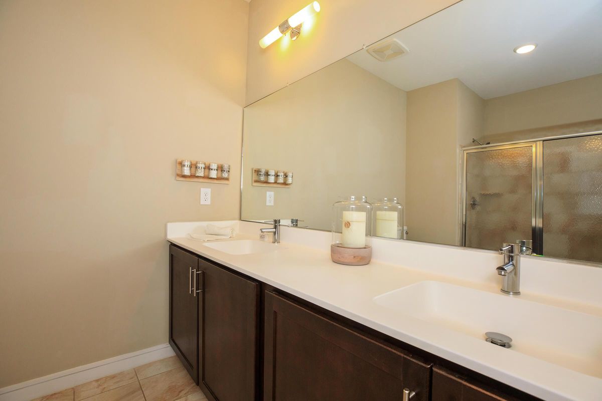 A spacious bathroom featuring a double sink countertop with dark wooden cabinetry, a large mirror above the sinks, a wall-mounted light fixture, and a shower area enclosed with glass doors. The decor is minimalistic with soft beige walls and a few decorative items on the countertop.