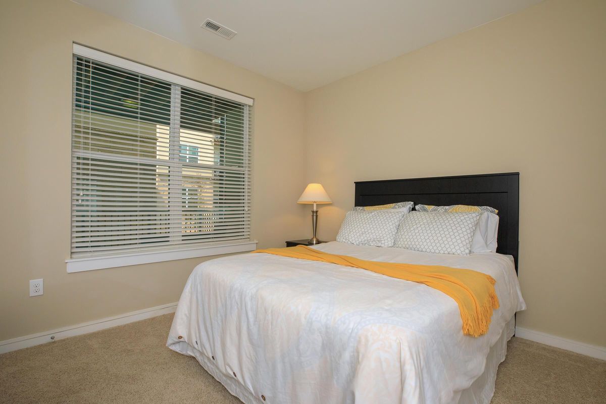 A cozy bedroom featuring a queen-sized bed with a white and yellow blanket, a window with horizontal blinds, and soft beige walls. There's a bedside lamp on a small table next to the bed, and the floor is carpeted. Natural light brightens the space, creating a warm and inviting atmosphere.