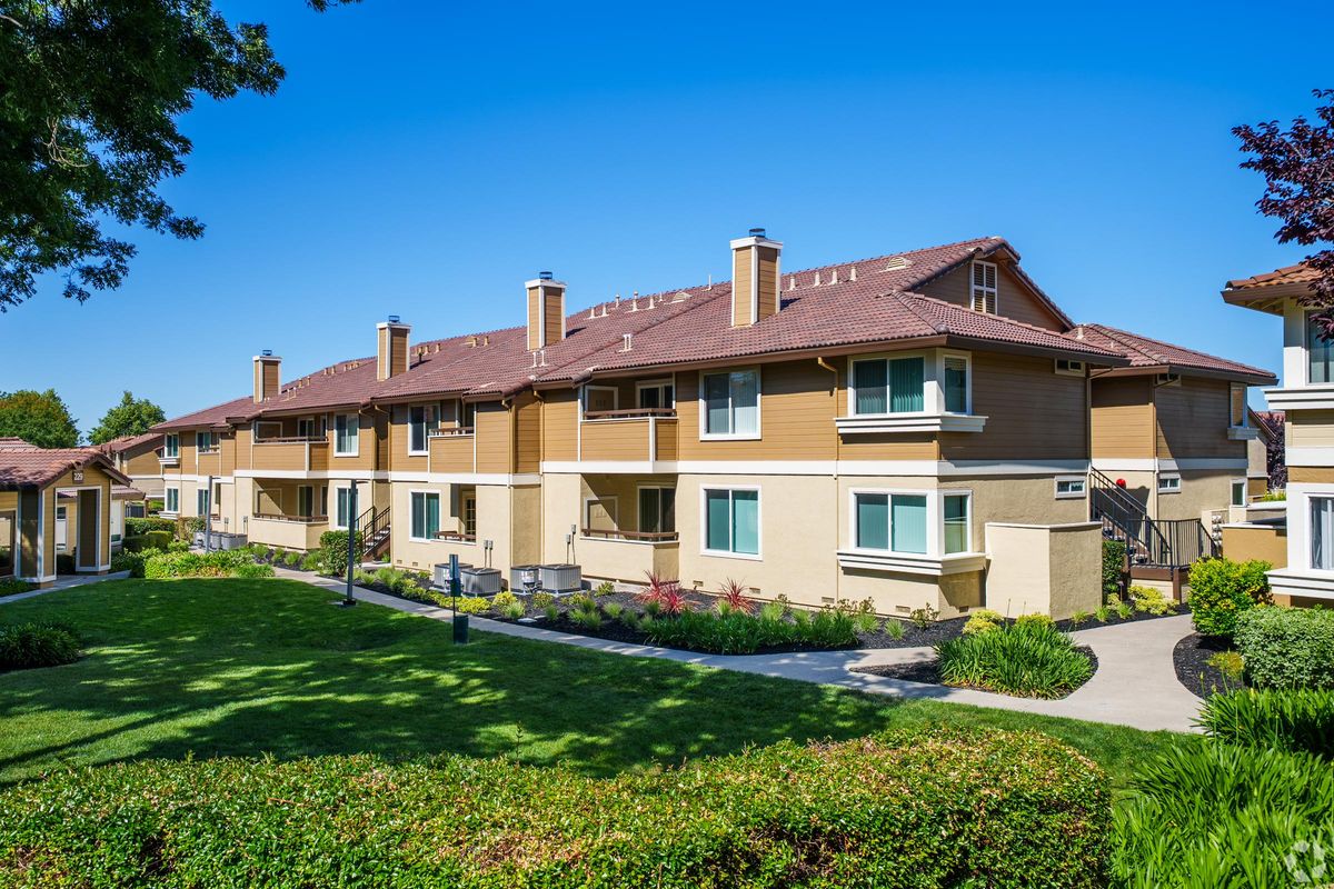 A row of modern two-story apartment buildings with a light brown exterior, surrounded by well-maintained green lawns and landscaped gardens. Clear blue skies above create a bright and inviting atmosphere, with a pathway leading through the greenery.