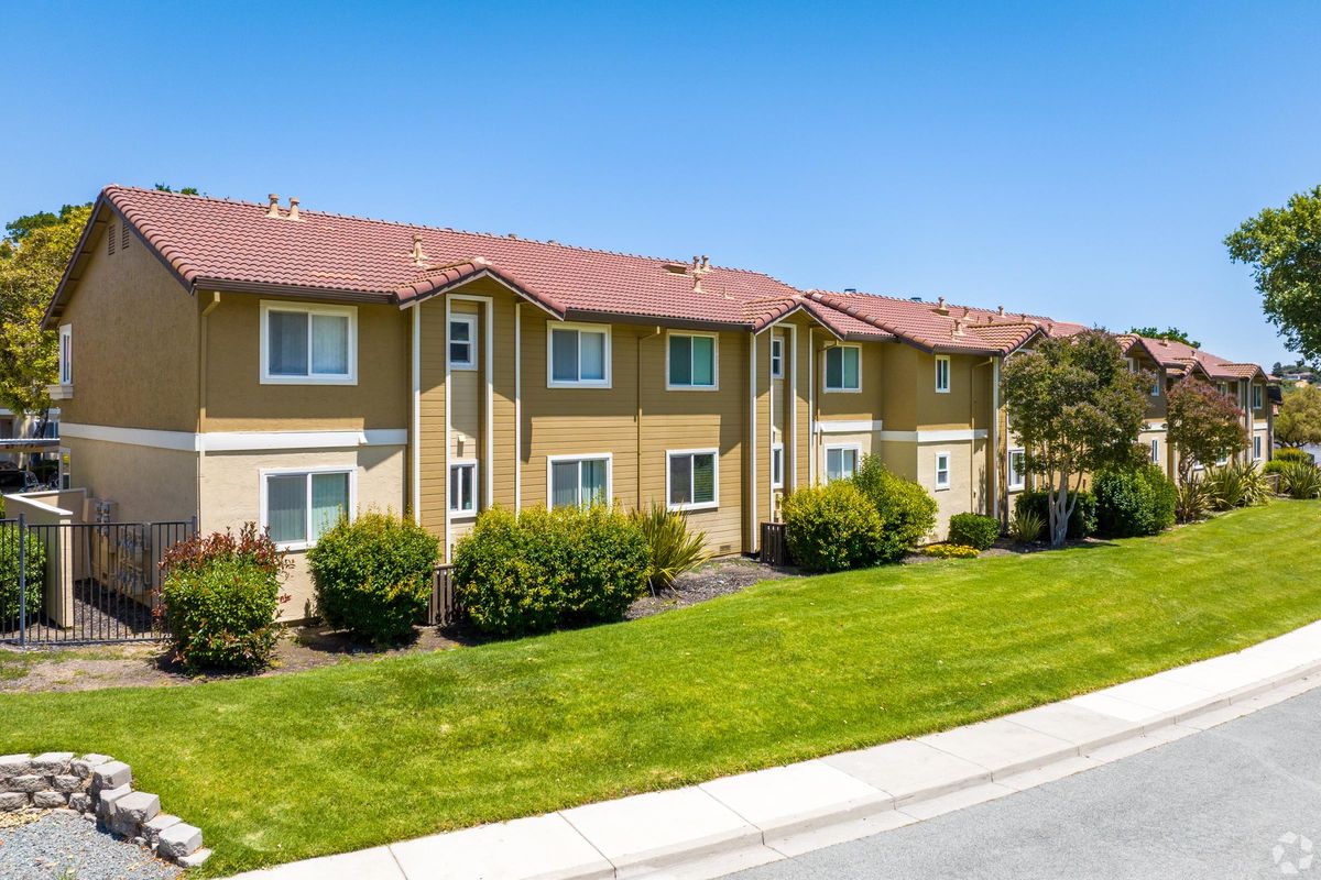 A row of residential buildings with two stories, featuring beige siding and red tile roofs. Lush green grass and shrubs surround the buildings, set against a clear blue sky. A paved road lines the front of the property, providing access to the homes.