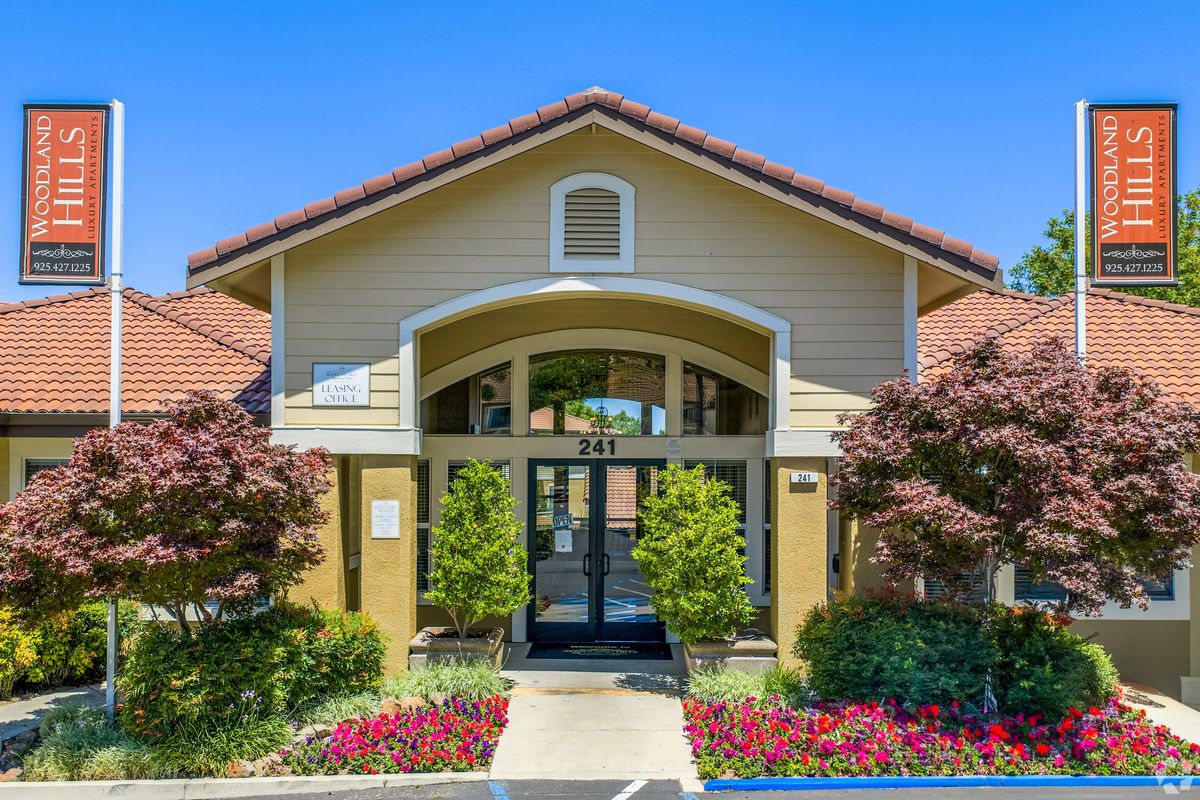 Exterior view of a building with a sloped roof and archway entrance. The building is labeled "Woodland Hills," with two signs on either side. There are colorful shrubs and flowers in front, along with well-maintained greenery and a clear blue sky in the background.