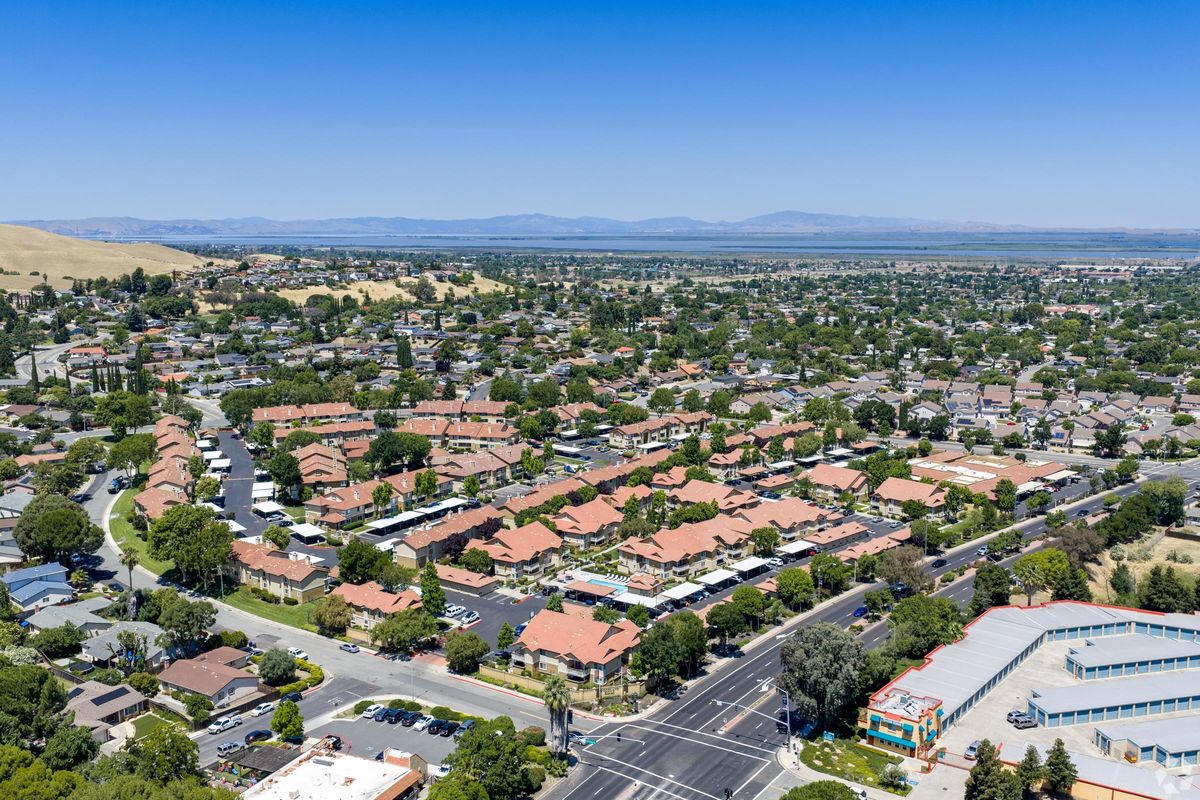 Aerial view of a suburban neighborhood featuring rows of houses with red roofs, green trees, and well-maintained streets. In the foreground, a road intersects with another, while in the background, rolling hills extend towards a blue sky and a distant view of mountains. The overall scene reflects a peaceful residential area.