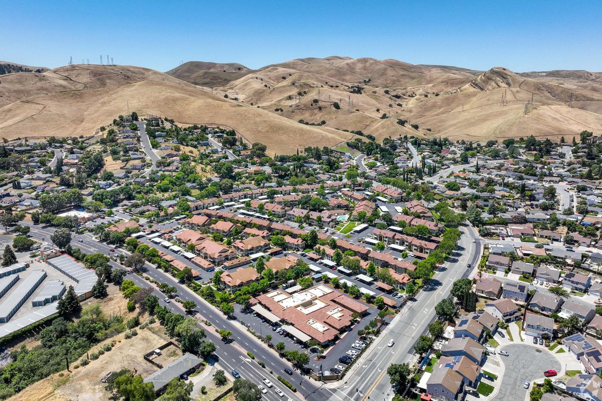 Aerial view of a suburban neighborhood with patches of green trees and brown hills in the background. The area features a mix of residential buildings and roads, with a clear blue sky overhead. The layout includes clusters of homes and some commercial structures adjacent to the main road.
