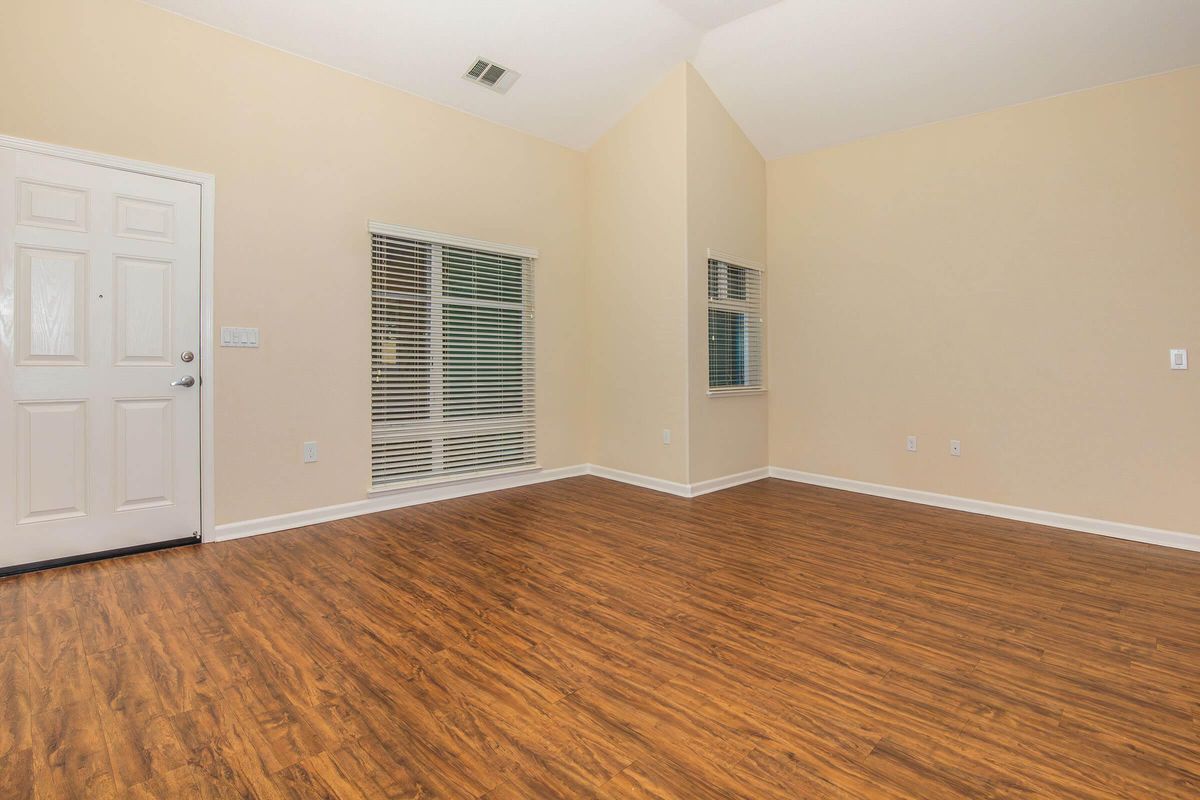 A spacious, empty room with light beige walls and a high ceiling. There is a white door on the left and large windows with horizontal blinds letting in natural light. The floor is made of polished, dark wood laminate, creating a warm and inviting atmosphere.