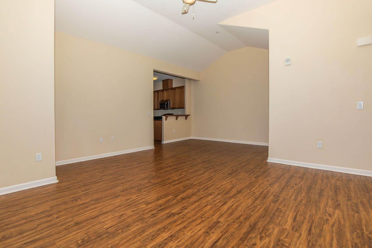 Spacious, empty room with light beige walls and a high ceiling. The flooring is a warm wood-like laminate. A doorway leads to a kitchenette area, which includes cabinets and appliances, partially visible in the background. The overall atmosphere is bright and open, ready for personalization.