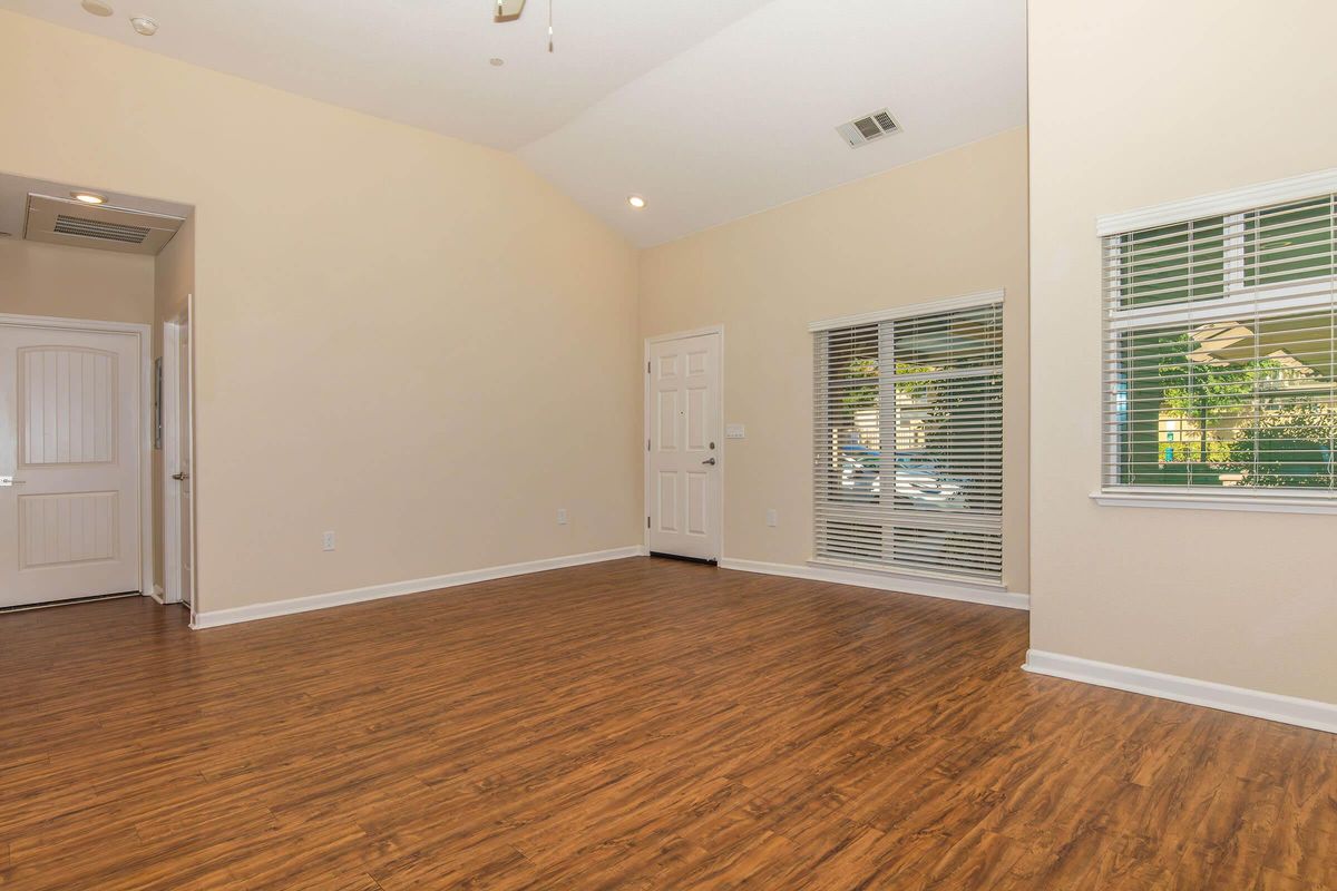 A spacious and well-lit interior of a living room featuring a vaulted ceiling, light beige walls, and wood-like flooring. There are green-tinted windows with white blinds, a front door, and a doorway on the left leading to another room. The area appears empty and ready for furnishing.