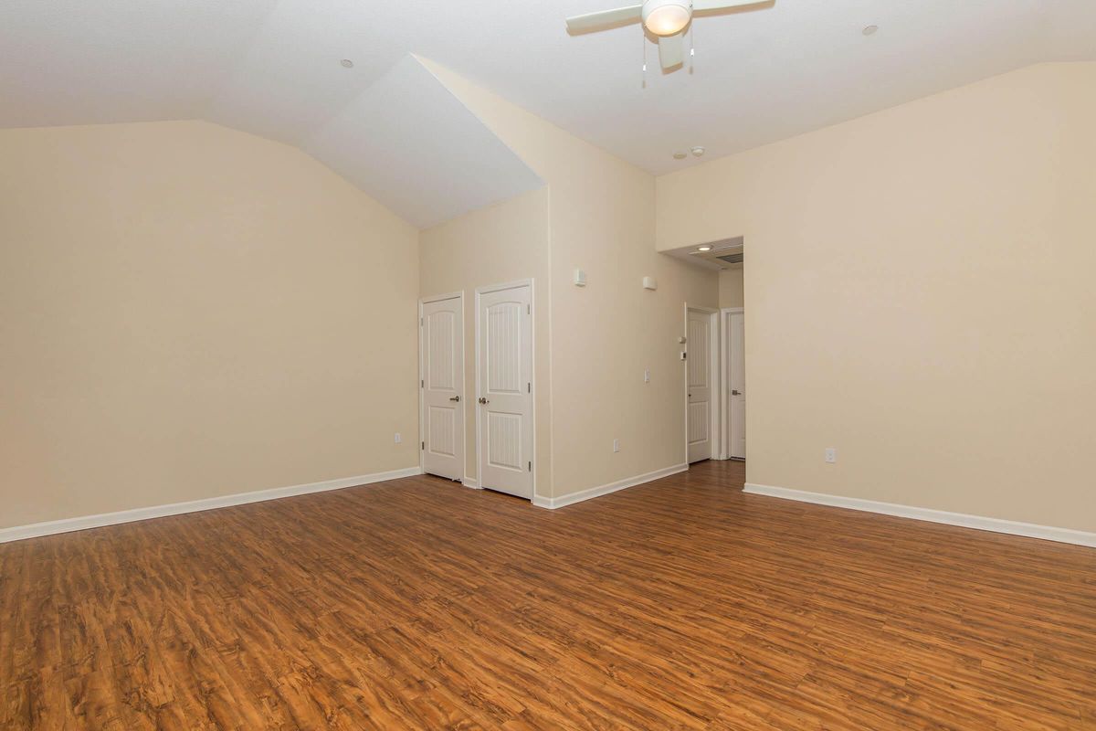Spacious interior room featuring light beige walls, high vaulted ceilings, and a faux wood floor. Two white double doors lead to adjacent spaces, while a ceiling fan is installed. The room is empty, providing a blank canvas for decoration and furniture arrangement. Natural light floods the space.