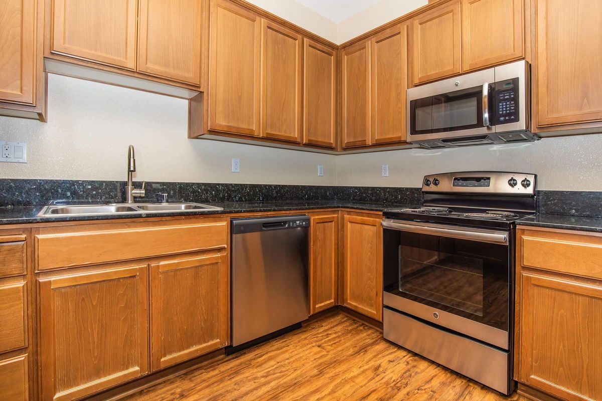A modern kitchen featuring wooden cabinets, a stainless steel refrigerator, a built-in dishwasher, an electric stove, and a microwave over the range. The countertops are dark with a subtle texture, and the floor is a warm-toned wood laminate. The space is well-lit and organized.