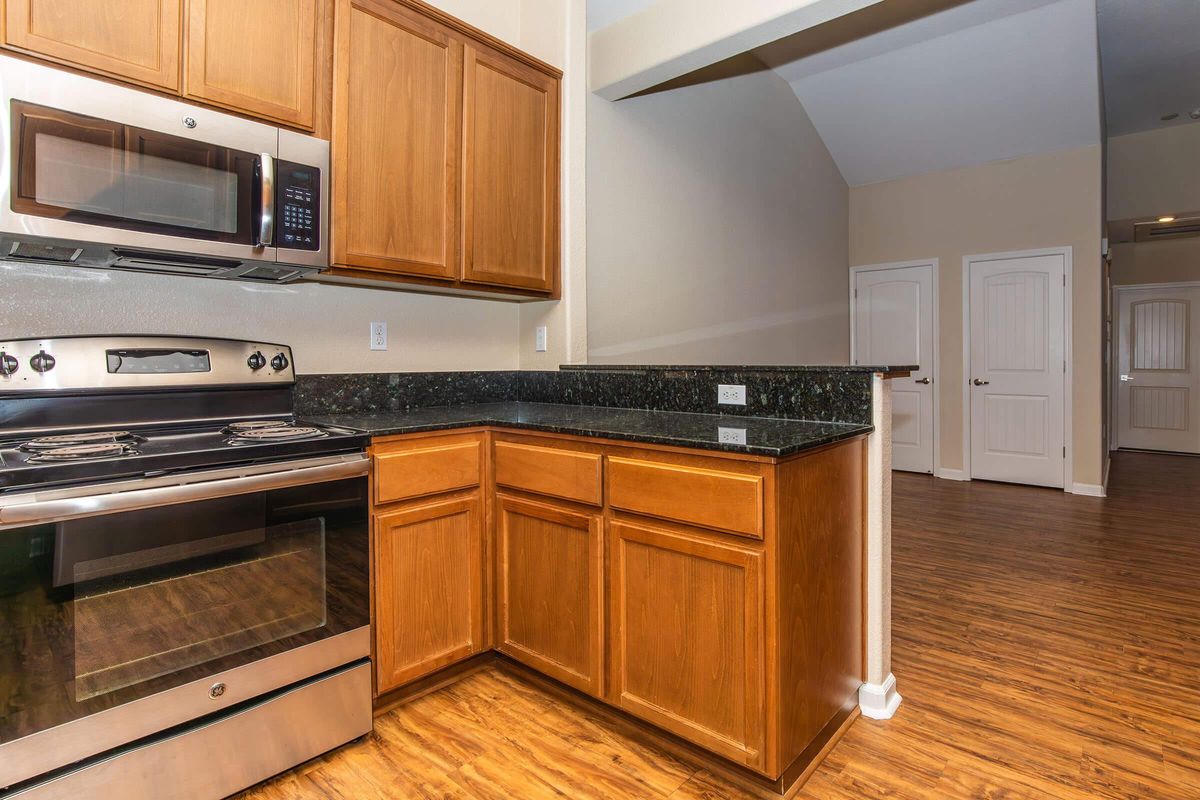Modern kitchen featuring wooden cabinets, a stainless steel oven, and a black granite countertop. The layout includes a view of a spacious area with light-colored walls. There are doors leading to other rooms, enhancing the open feel of the space.
