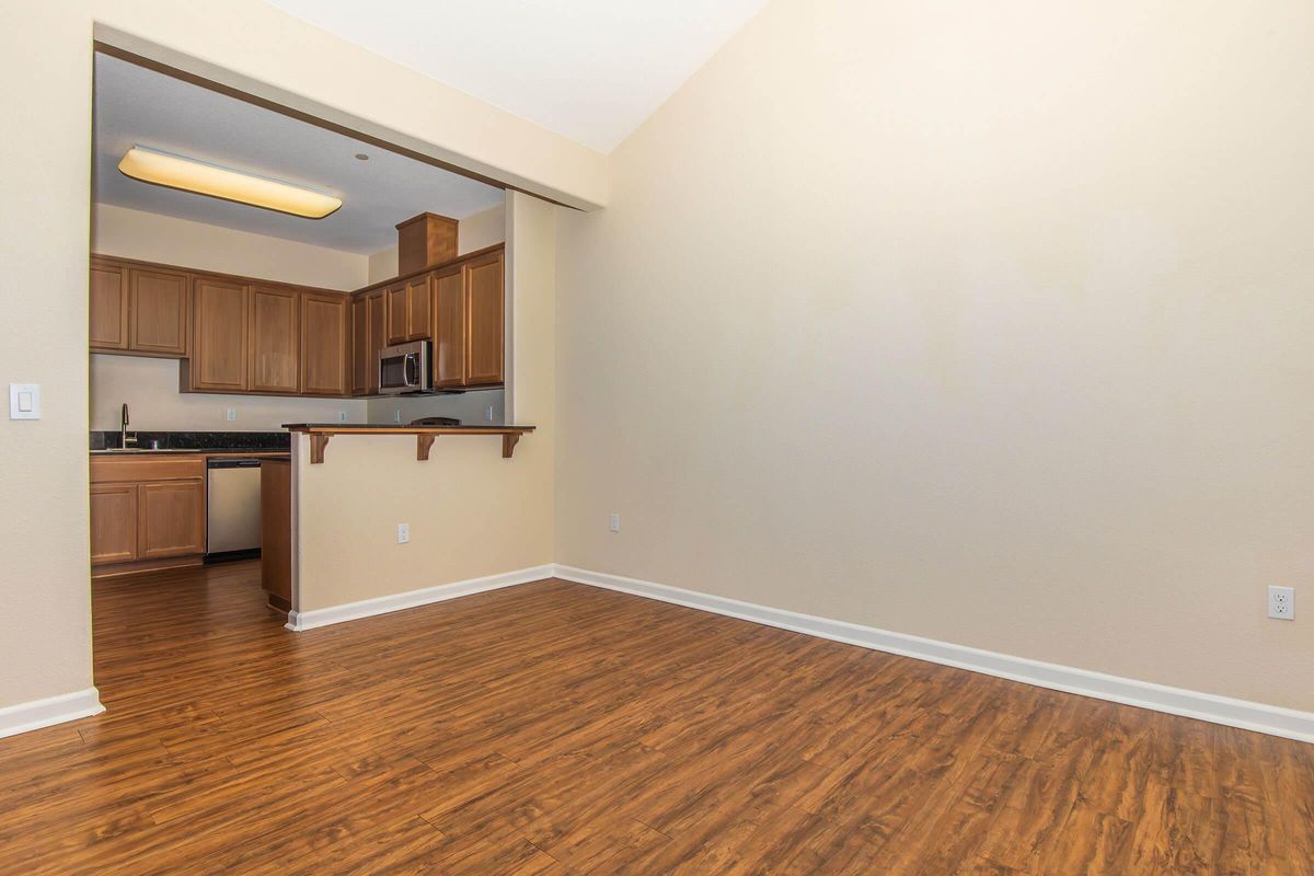 A stylish, spacious interior featuring a living area with laminate flooring and a light-colored wall. In the background, a modern kitchen with wooden cabinetry, stainless steel appliances, and a breakfast bar is visible. The space is bright and inviting, perfect for home decor or customization.