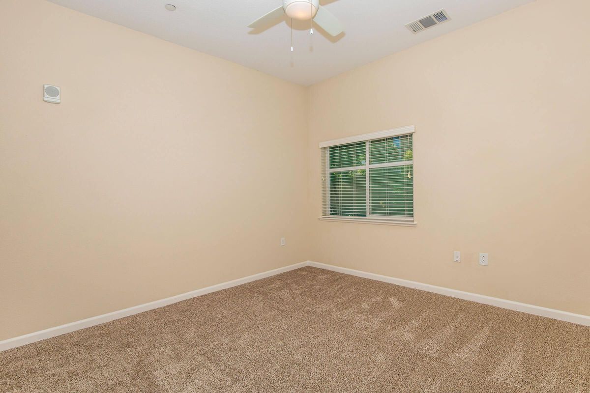 A spacious, empty room with light beige walls and carpet. A ceiling fan is mounted in the center, and there is a window with white blinds allowing natural light to enter. The room features a small air vent and two electrical outlets on one wall.