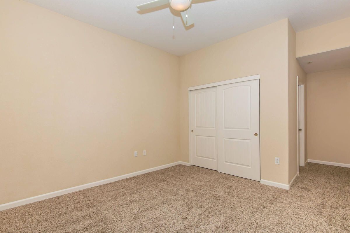 A bright, empty room with beige walls and light brown carpet. There is a ceiling fan in the center and a pair of white double doors leading to a closet. A doorway on the right leads to another room. The space is well-lit and features a clean, minimalist design.