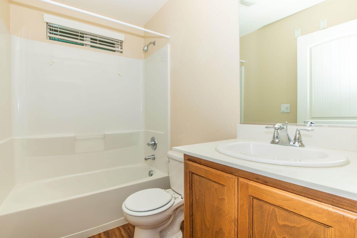 A clean, modern bathroom featuring a white bathtub with a shower, a sink with a wooden cabinet underneath, and a large mirror. The walls are painted a light color, and there is a window above the bathtub for natural light. The flooring is wood-like, adding warmth to the space.
