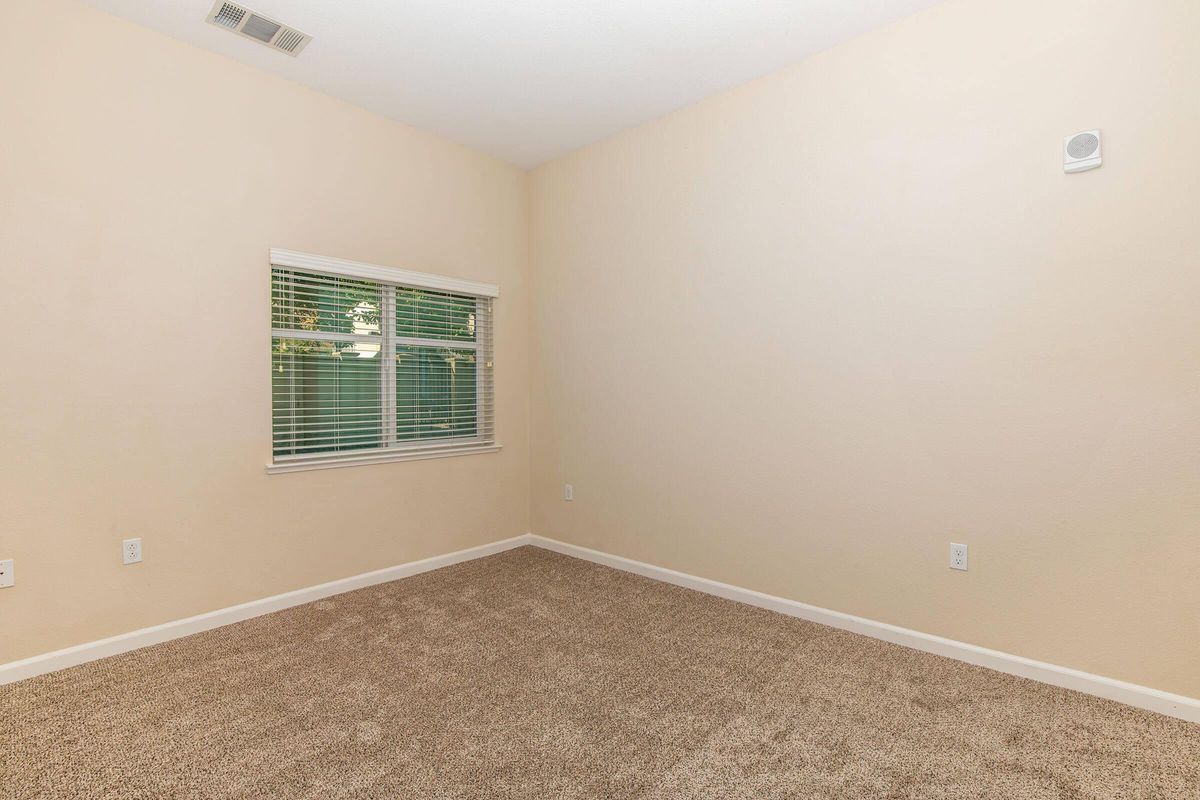 Empty room with beige walls and carpeted floor. A window with white blinds allows natural light to enter. The room is unfurnished, with electrical outlets visible on the walls. A small vent is located near the ceiling, and there is a sense of openness and simplicity in the design.