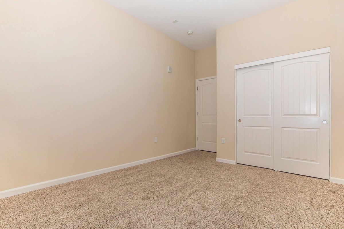 A vacant room with beige walls and a light carpeted floor. There are two doors: one is a closet with white double doors, and the other leads to another space. The room is well-lit and has minimal decoration, creating a spacious and open feel.