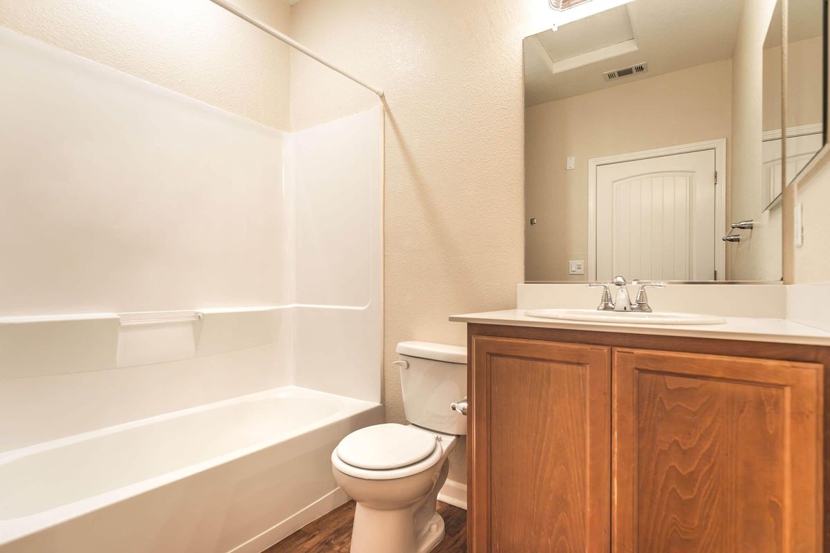 A clean, modern bathroom featuring a white bathtub with a shower, a toilet, and a wooden vanity with a sink. The walls are a light beige, and there is a large mirror above the sink. The floor has a wood-like finish, and a door is visible in the background.