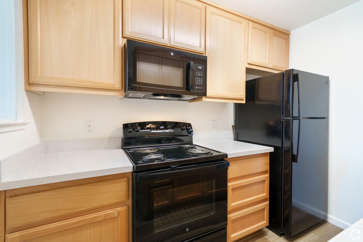 A modern kitchen featuring light wood cabinets, a black stove and microwave, and a black refrigerator. There is a white countertop, and the overall design is clean and contemporary with natural lighting.