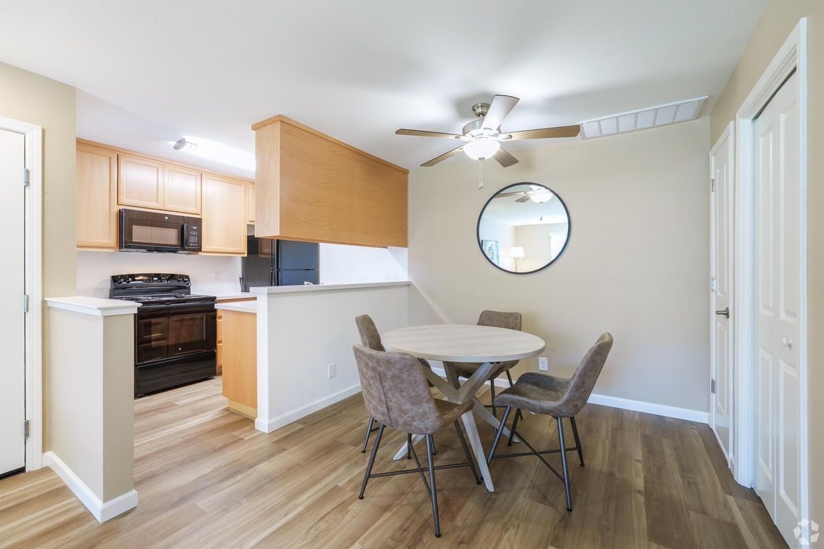 A modern kitchen and dining area featuring light wood cabinets, a round table with four gray chairs, and a ceiling fan. The kitchen includes a black stove and microwave. A circular mirror is on the wall, and the flooring is light-colored laminate, creating a clean and inviting atmosphere.