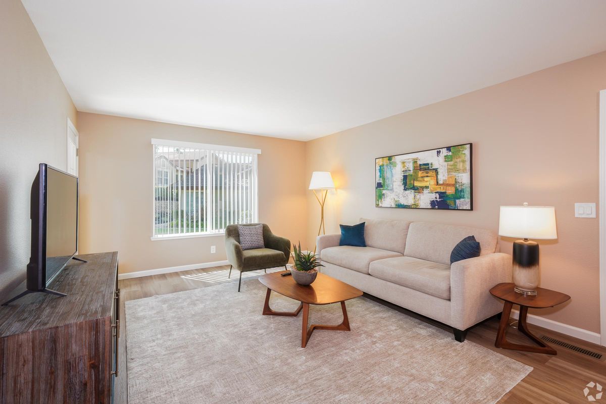 A cozy living room featuring a light-colored couch, a green accent chair, and a coffee table. There's a television on a wooden console, a lamp providing warm light, and a piece of abstract art on the wall. Natural light streams through a window with blinds. The floor is light wood, and a soft area rug is under the coffee table.