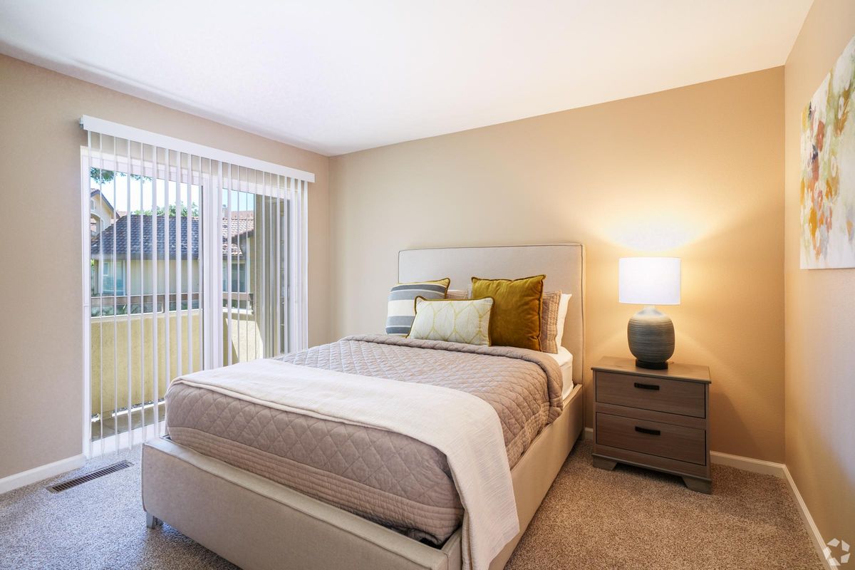 Bright and cozy bedroom featuring a queen-sized bed with decorative pillows, a nightstand with a lamp, and a window with vertical blinds allowing natural light. Soft beige walls and carpet create a warm atmosphere. A piece of decorative art hangs on the wall, enhancing the room's aesthetic.