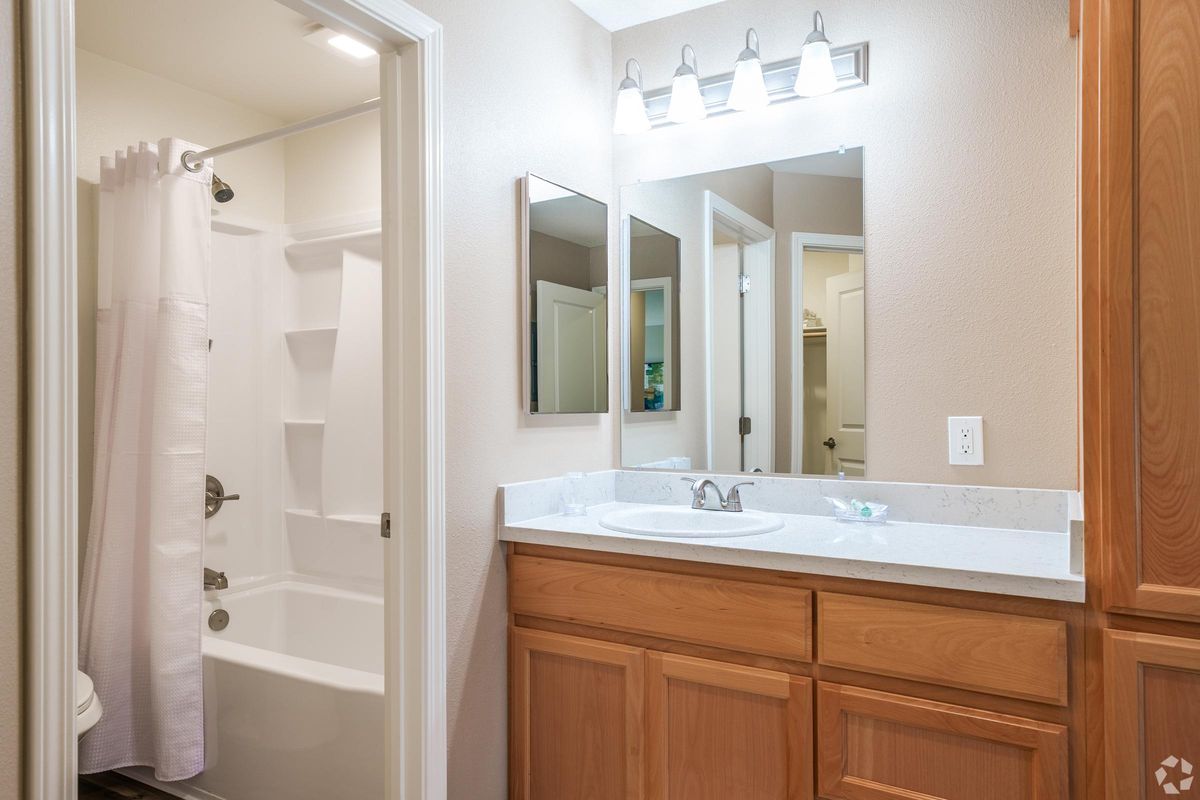 A well-lit bathroom featuring a white bathtub with a shower curtain, a wooden vanity with a sink, and a large mirror above it. The walls are painted a light color, and there is a modern light fixture with multiple bulbs. A door leads to another room in the background.